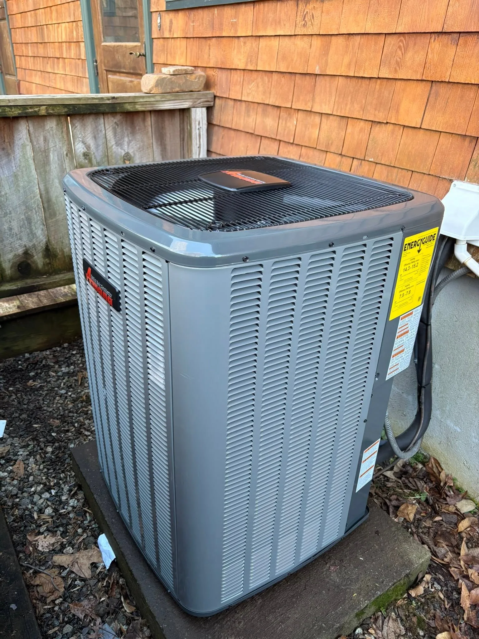 A new Amana condenser installed on a concrete pad beside a cedar shingle house, the EnergyGuide label still on the cabinet and the electrical whip run neatly into the disconnect.