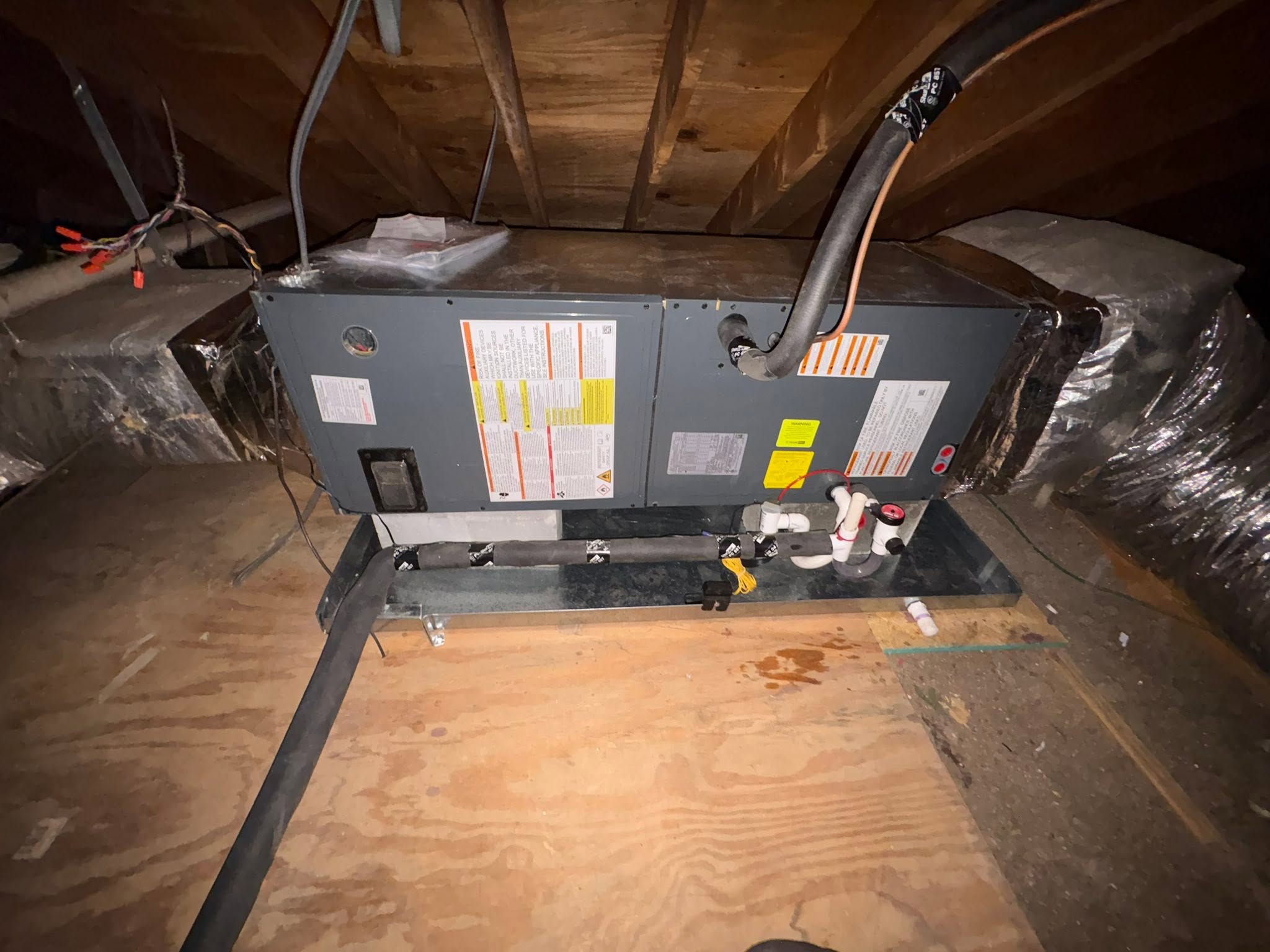 An attic air handler on a plywood platform with an insulated refrigerant line and a secondary drain pan.